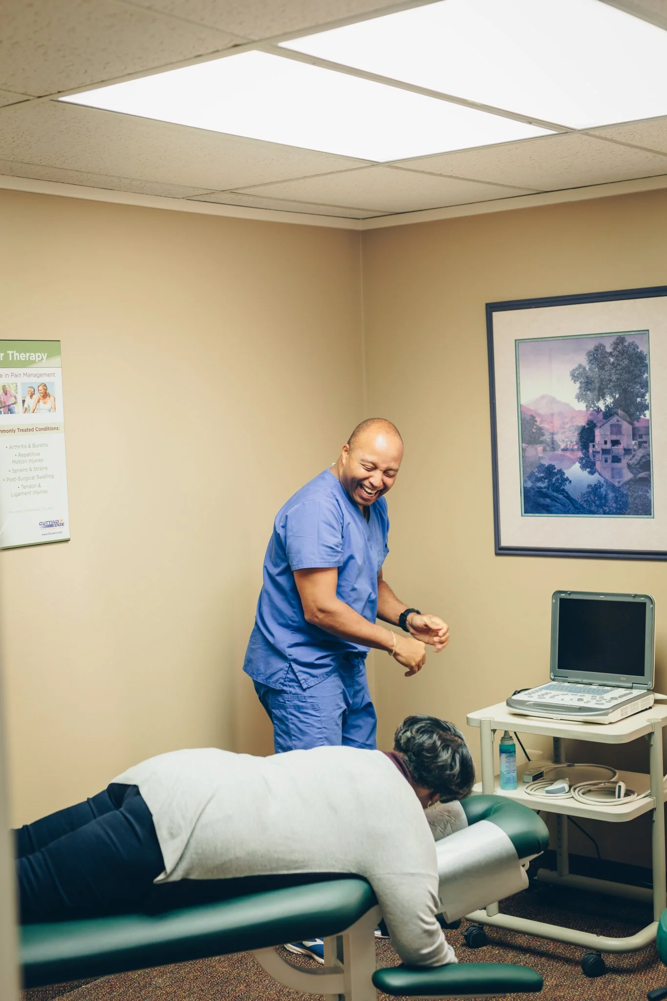 Provider in blue scrubs performing a physical examination on a patient lying on a treatment table in a clinical room